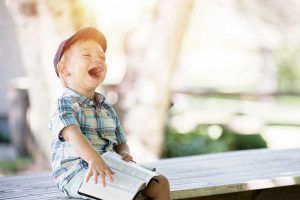 Un niño pequeño con gorra riendo a carcajadas mientras está sentado con un libro en sus manos. Terapeuta ocupacional infantil.