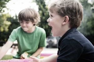 Dos niños jugando al aire libre; uno de ellos, con camiseta verde, está difuminado en el fondo, mientras que el otro, con camiseta negra, está gritando con expresión de sorpresa o alegría. Terapeuta ocupacional infantil.