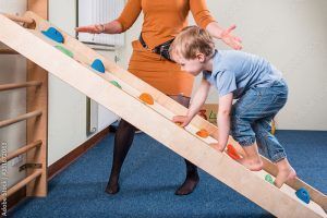 Niño pequeño escalando una rampa de madera con agarraderas de colores mientras una mujer en vestido naranja le guía. ¿Por qué es tan importante en los niños y las niñas la integración sensorial? ¿Qué es la integración sensorial?