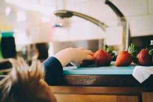 La imagen muestra la mano de un niño pequeño alcanzando una fresa fresca sobre una tabla de cortar en la cocina, ilustrando el descubrimiento y la curiosidad, con un enfoque suave y un fondo de cocina borroso. Mi hijo solo come yogures, pasta, patatas…