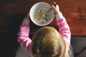 La imagen captura una vista aérea de una niña pequeña en pijama comiendo cereal con leche en un tazón blanco, sosteniendo una cuchara, destacando la independencia durante la hora del desayuno en un entorno hogareño. Mi hijo solo come yogures, pasta, patatas…