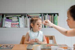 La imagen muestra a una niña sonriente sentada a la mesa con un babero, esperando comer una cucharada de comida de la mano de un adulto, en un ambiente familiar con estantes de libros al fondo. Mi hijo solo come yogures, pasta, patatas…