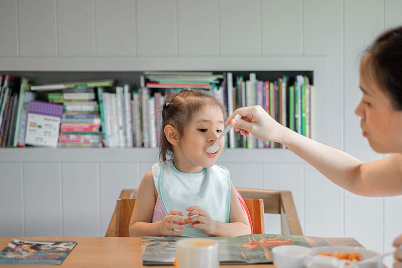 La imagen muestra a una niña sonriente sentada a la mesa con un babero, esperando comer una cucharada de comida de la mano de un adulto, en un ambiente familiar con estantes de libros al fondo. Mi hijo solo come yogures, pasta, patatas…