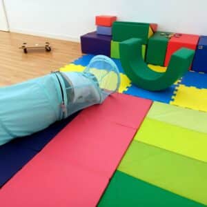 Una zona de juegos infantil con colchonetas de espuma de colores, un túnel de juegos, bloques blandos y una tabla de skate sobre un suelo de madera; todo ello dispuesto cuidadosamente para seguir las recomendaciones de una terapeuta ocupacional infantil.