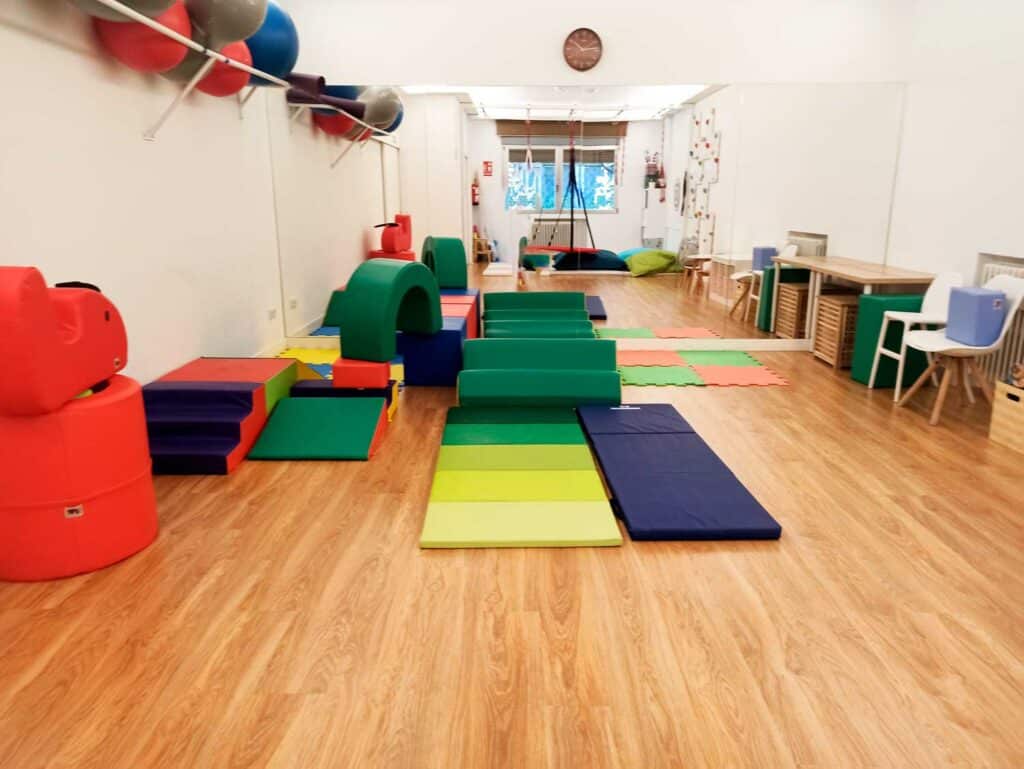 Sala de juegos interior diseñada por una terapeuta ocupacional infantil, con bloques de espuma para trepar, colchonetas acolchadas y material lúdico de colores sobre un suelo de madera. Las paredes están decoradas con estanterías, mesas y pelotas de ejercicio.