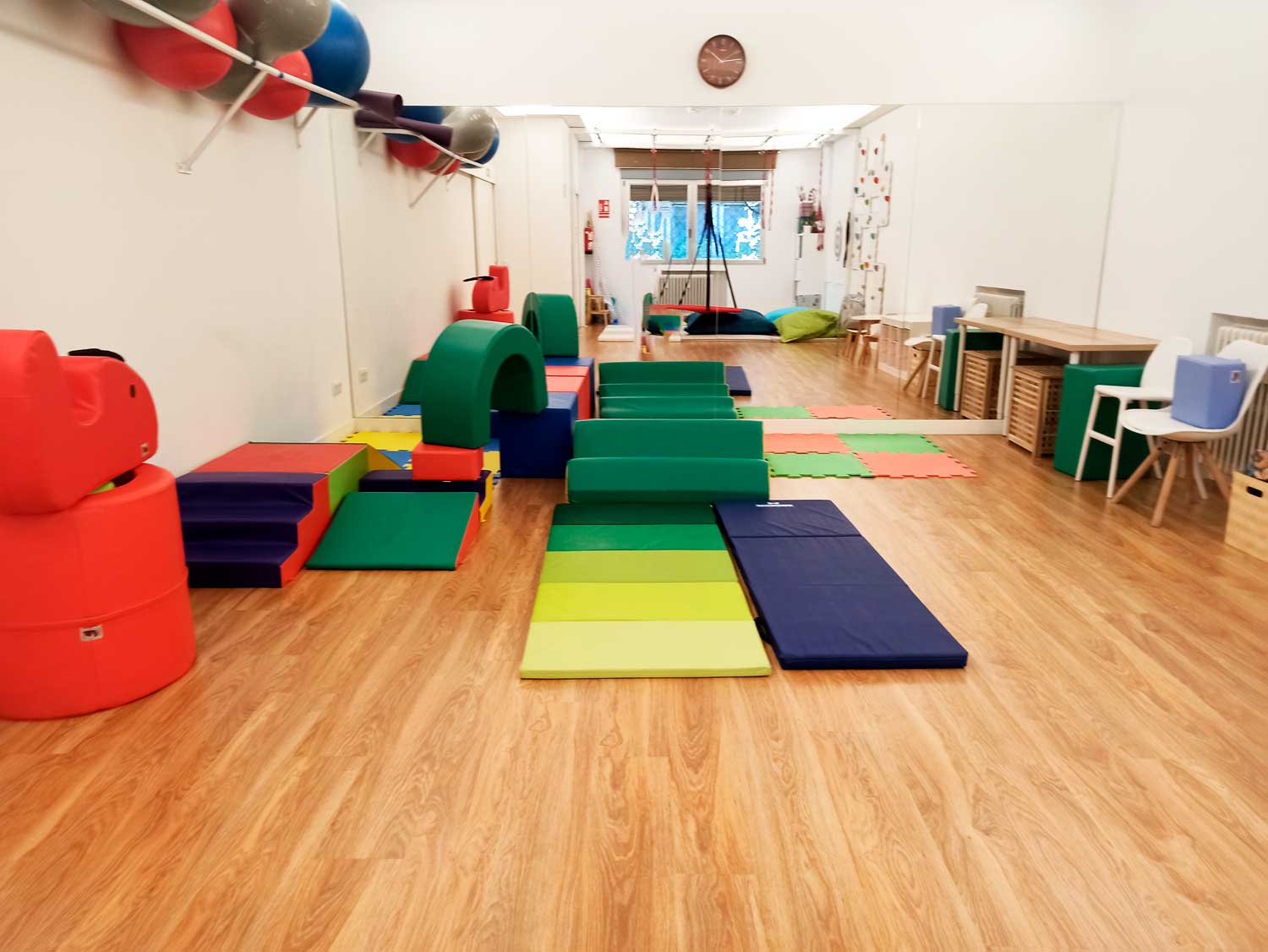 Sala de juegos interior diseñada por una terapeuta ocupacional infantil, con bloques de espuma para trepar, colchonetas acolchadas y material lúdico de colores sobre un suelo de madera. Las paredes están decoradas con estanterías, mesas y pelotas de ejercicio.