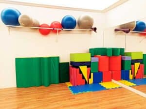 Las coloridas colchonetas de espuma y las pelotas de ejercicio están apiladas ordenadamente en una sala con una pared de espejos y suelo de madera, creando un ambiente acogedor que suele utilizar una terapeuta ocupacional infantil para fomentar el desarrollo de los niños a través del juego y el movimiento.