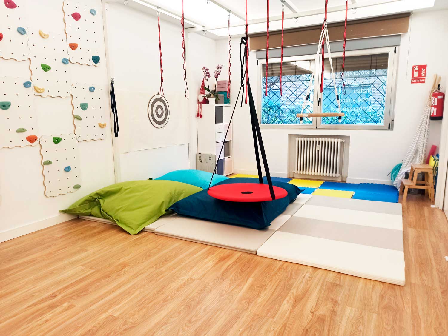 Sala de juegos sensoriales con colchonetas acolchadas, columpios colgantes, cojines grandes, paneles de rocódromo y una ventana con rejas. El suelo es de madera y hay un extintor en la pared. Diseñado para acoger sesiones dirigidas por una terapeuta ocupacional infantil, este espacio anima a los niños a explorar y desarrollar sus habilidades sensoriales y motoras en un entorno seguro.