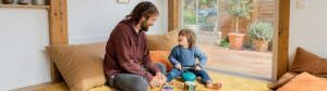 Un adulto y un niño están sentados en el suelo, en el interior de una casa, tocando instrumentos musicales y sonriéndose el uno al otro, lo que ilustra un momento alegre que pone de relieve qué es la atención temprana. A su alrededor hay cojines grandes, y una ventana al fondo deja entrever un patio lleno de plantas, lo que crea un ambiente cálido y acogedor.