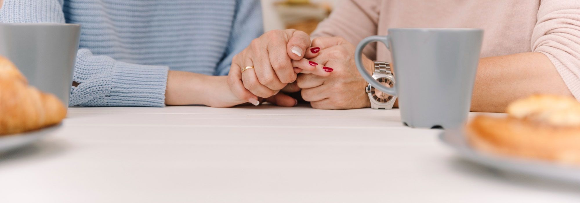 Dos personas están sentadas a una mesa, cogidas de la mano, con tazas y platos de bollería delante de ellas. Solo se les ven los brazos y las manos.