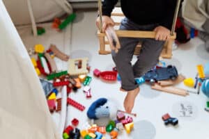 Un niño está sentado en un columpio de madera sobre una alfombra de juegos en la que hay esparcidos bloques de construcción de colores, peluches y otros juguetes, lo que crea un entorno acogedor que refleja el ambiente estimulante de la terapia ocupacional infantil en Madrid.