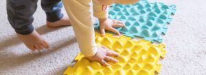 Un bebé en pijama toca unas alfombras sensoriales con texturas sobre un suelo alfombrado, explorando la integración sensorial con una alfombra amarilla y otra verde azulado.