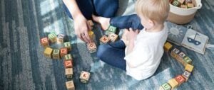 Un adulto y un niño pequeño están sentados sobre una alfombra azul, jugando con bloques de madera con letras del abecedario. Cerca de ellos hay una cesta con bloques y un libro de páginas blandas, lo que crea el ambiente ideal para una actividad significativa sobre mí mientras exploran las letras juntos.