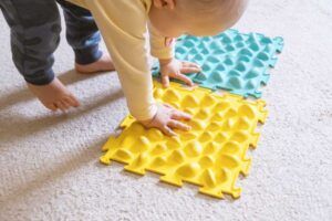 Un niño realiza ejercicios de integración sensorial tocando unas alfombrillas de espuma con texturas que encajan entre sí, una amarilla y otra verde azulada, mientras está arrodillado sobre un suelo alfombrado.