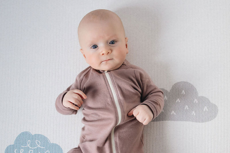 Un bebé con un pijama malva con cremallera, tumbado sobre una colchoneta con un estampado de nubes. Qué es la edad corregida.